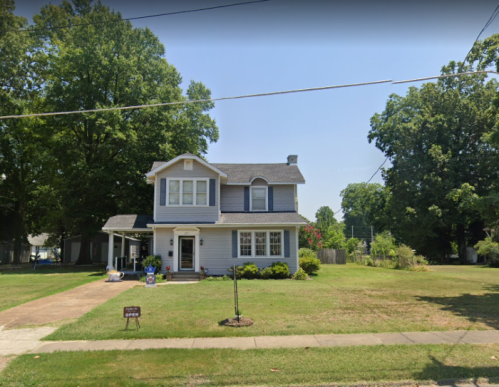 A two-story gray house with a front yard, surrounded by trees, on a sunny day. A sign is visible in the yard.