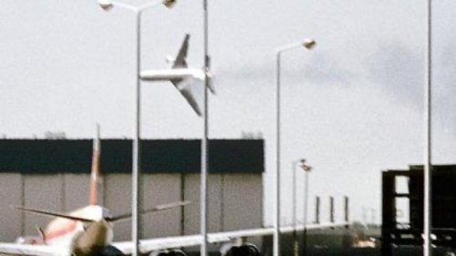A jet aircraft takes off near an airport, with another plane visible on the tarmac and smoke in the background.