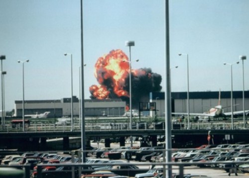 Explosion at an airport, with flames and smoke rising behind parked cars and aircraft.
