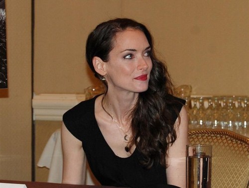 A woman with long, wavy hair and red lipstick sits at a table, looking to the side with a thoughtful expression.