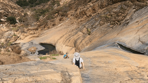 Two people climbing a rocky slope near a small pool of water, surrounded by rugged terrain and greenery.