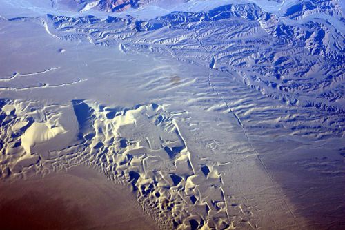 Aerial view of a vast, textured landscape featuring snow and ice formations, with ridges and valleys visible.