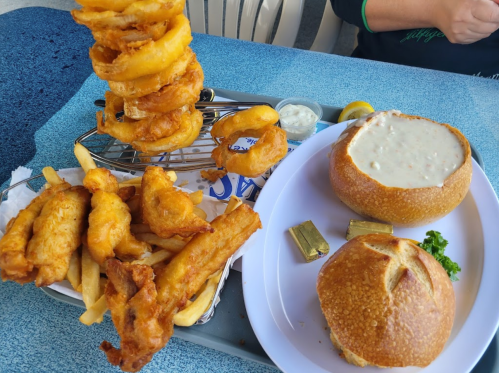 A tray with onion rings, fried fish, fries, and a bread bowl filled with clam chowder, garnished with lemon.