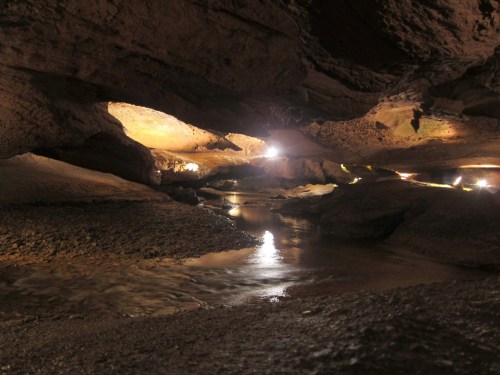 A dimly lit cave interior with a winding stream and rocky formations, illuminated by soft lights.