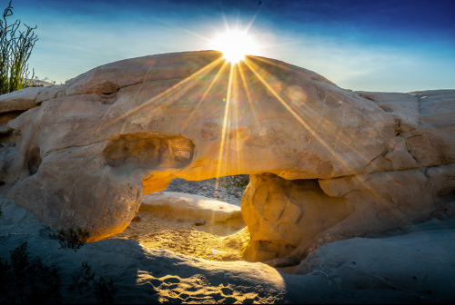 A sunburst shines through a natural rock arch, illuminating sandy ground beneath in a serene landscape.