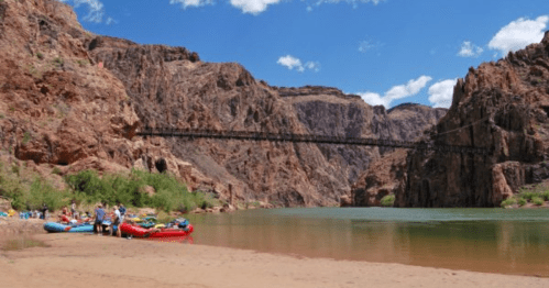 A sandy riverbank with colorful rafts, surrounded by towering red rock cliffs and a bridge in the background under a blue sky.