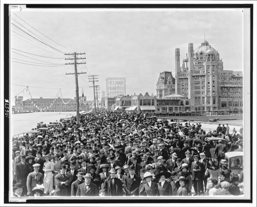 A large crowd of people in early 20th-century attire gathered along a waterfront promenade with buildings in the background.