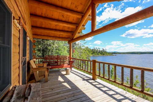 A wooden cabin porch overlooking a serene lake, surrounded by trees and a bright blue sky with fluffy clouds.