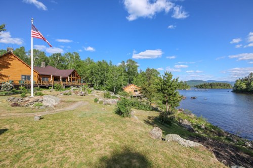 A scenic view of a lakeside cabin with an American flag, surrounded by trees and rocky shoreline under a blue sky.