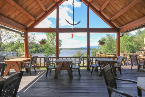 A cozy wooden patio with tables and chairs overlooking a serene lake and trees under a bright blue sky.