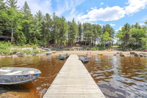 A wooden dock extends over a calm lake, surrounded by trees and a sandy beach with boats in the background.