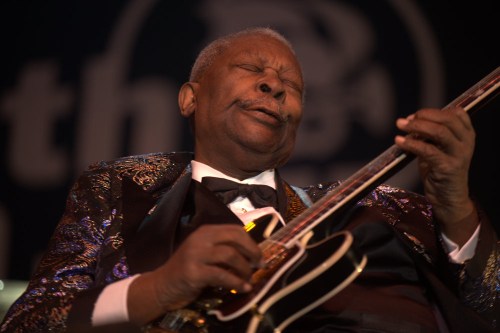 A musician passionately plays guitar on stage, wearing a shiny suit and expressing deep emotion through his performance.