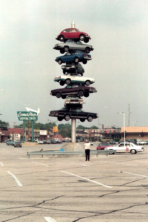 A tower of stacked cars in a parking lot, with a person taking a photo in the foreground.