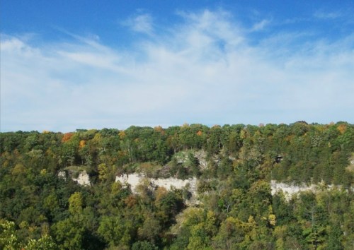 A scenic view of a forested cliff under a blue sky with scattered clouds and hints of autumn foliage.