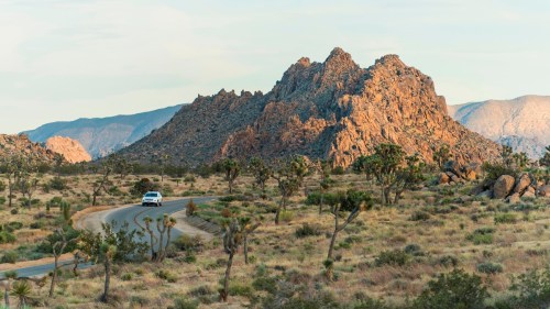A car drives along a winding road through a desert landscape with rocky mountains and sparse vegetation.