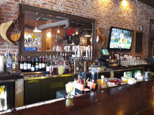 A bar with a brick wall, shelves of liquor, a TV showing a sports game, and a wooden counter with drinks and utensils.