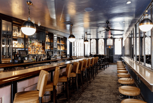 A stylish bar interior featuring a long counter, high stools, and shelves stocked with colorful bottles.