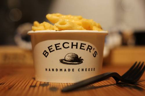 A cup of creamy macaroni and cheese from Beecher's Handmade Cheese, with a black fork beside it on a wooden table.