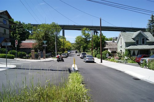 A quiet street lined with houses, greenery, and parked cars, with a bridge overhead in the background.