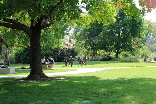 A sunny park scene featuring a tree, grassy area, and a group of children sculptures playing together.