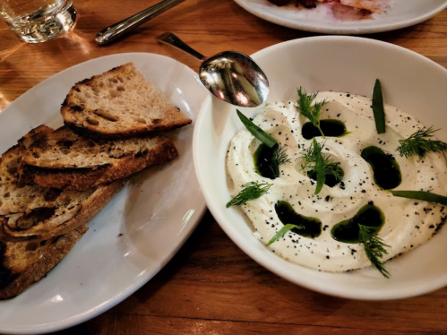 A bowl of creamy dip garnished with herbs and olive oil, served with toasted bread on a wooden table.