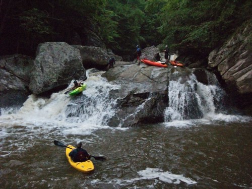 Kayakers navigate rocky rapids, with one paddler in a yellow kayak and others on the rocks in the background.