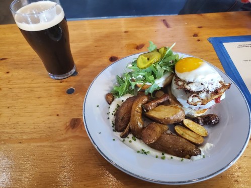A plate with a burger topped with a fried egg, roasted potatoes, and a side salad, next to a glass of dark beer.