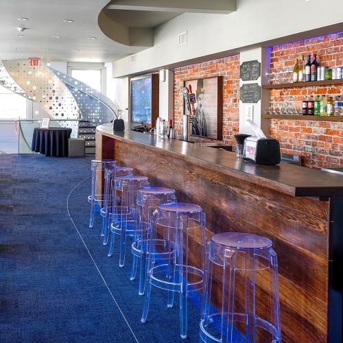 A modern bar with a wooden counter, clear stools, and a brick wall, featuring shelves of drinks and a spiral staircase.