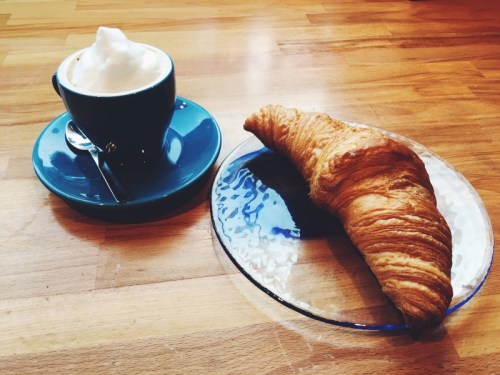 A blue cup of cappuccino beside a flaky croissant on a glass plate, set on a wooden table.