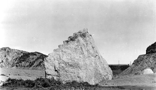A large, jagged rock formation stands prominently in a barren landscape, with hills in the background and a clear sky.
