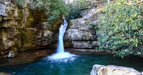 A serene waterfall cascades over rocky cliffs into a tranquil pool, surrounded by lush greenery.
