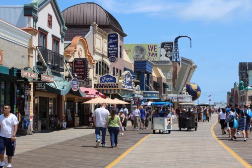 A bustling boardwalk scene with shops, people walking, and colorful signage under a clear blue sky.