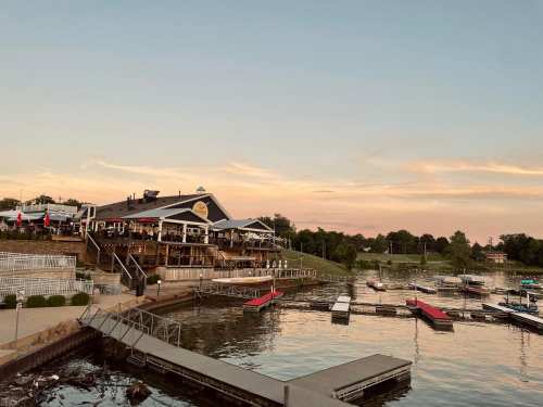 A lakeside restaurant at sunset, with boats docked and a scenic view of the water and surrounding greenery.