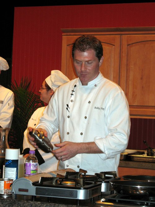 A chef in a white coat prepares food at a cooking demonstration, with kitchen equipment and assistants in the background.