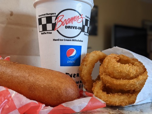 A corndog, onion rings, and a Pepsi cup from Boomer's Drive-In on a checkered paper background.