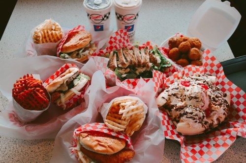 A variety of fast food items including burgers, fries, chicken, and desserts, served in red and white checkered paper.