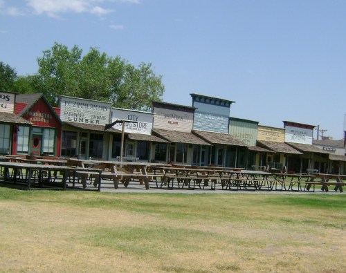 A row of old western-style buildings with wooden benches in front, set against a clear blue sky.