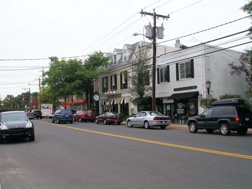 A street scene featuring parked cars, shops, and trees, with power lines overhead on a cloudy day.