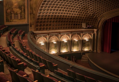 Interior of a vintage theater featuring ornate architecture, empty seats, and a stage with red curtains.