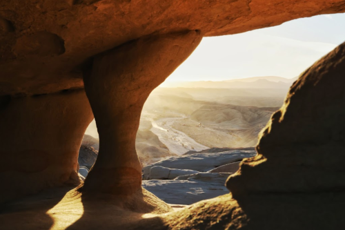 Sunlight streams through a rock formation, illuminating a desert landscape with soft hills in the background.