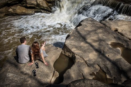 A couple sits on a rock by a waterfall, enjoying the view and the sound of flowing water.