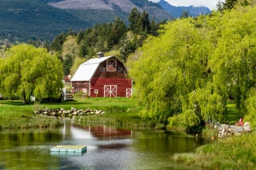 A red barn beside a calm pond, surrounded by lush green trees and mountains in the background on a sunny day.
