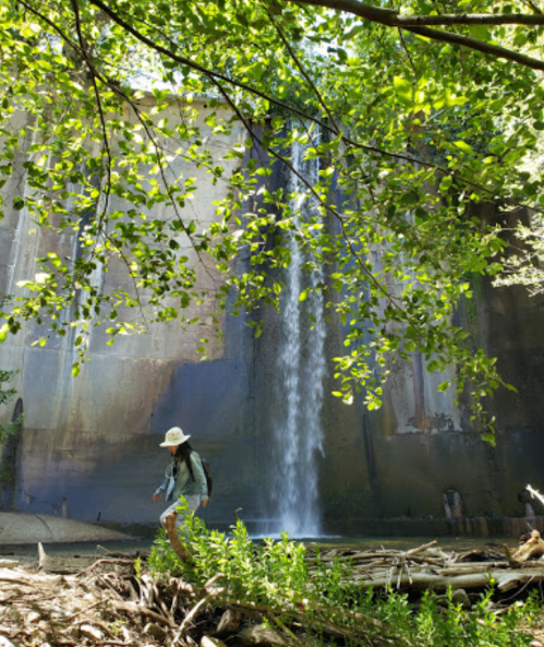 A person in a hat walks near a waterfall surrounded by lush greenery and a concrete wall.