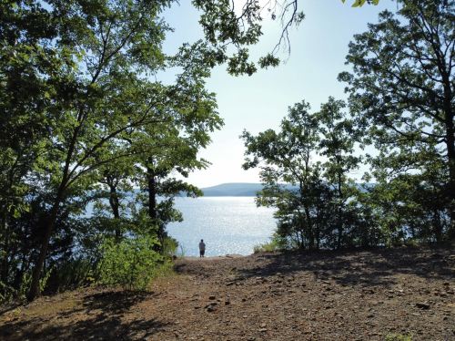 A serene lakeside view framed by trees, with a person standing near the water's edge under a clear blue sky.