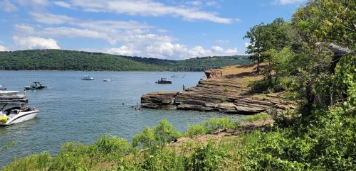A scenic view of a lake with boats, rocky shore, and lush green hills under a blue sky with clouds.