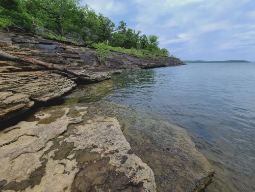 Rocky shoreline with clear water and lush greenery under a cloudy sky.