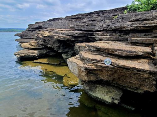 Rocky shoreline with layered stones beside calm water, reflecting a cloudy sky. A small circular object rests on the rocks.