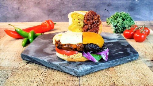 A gourmet burger with cheese, tomatoes, onions, and jalapeños on a slate platter, surrounded by fresh vegetables.