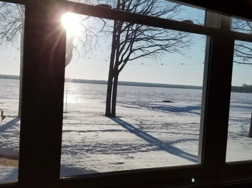 Sunlight streams through a window, illuminating a snowy landscape with trees and a frozen lake in the distance.
