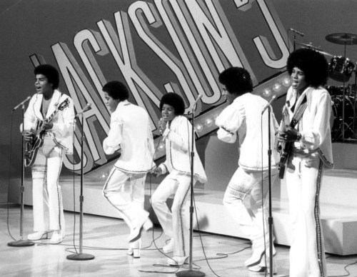 The Jackson 5 perform on stage in matching white outfits, singing and playing instruments in a lively black-and-white scene.
